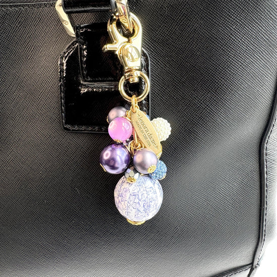 Beaded bag charm with a mix of bead styles in shades of purple, blue, gold and pearl. One larger round bead in a purple and white toile print hangs at the bottom and a gold trigger clasp on top of all of the beads. Brass oval hang tag near the trigger clasp that says "Lenora Dame Handmade in USA". Lenora Dame bag charm.