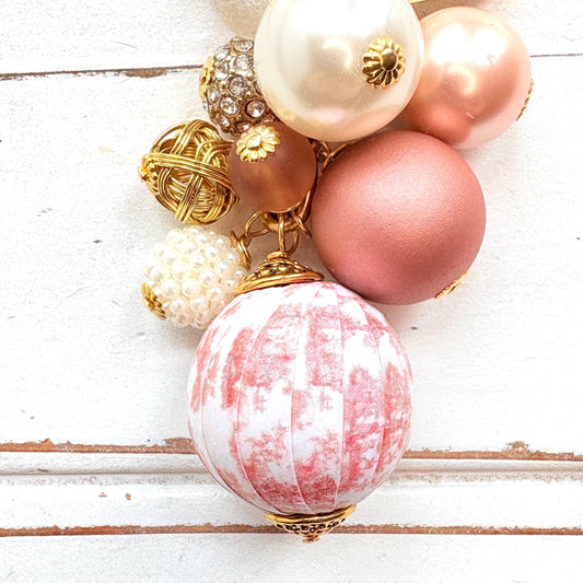 Beaded bag charm with a mix of bead styles in shades of pink, peach, gold and pearl. One larger round bead in a pink and white toile print hangs at the bottom and a gold trigger clasp on top of all of the beads. Brass oval hang tag near the trigger clasp that says "Lenora Dame Handmade in USA". Lenora Dame bag charm.