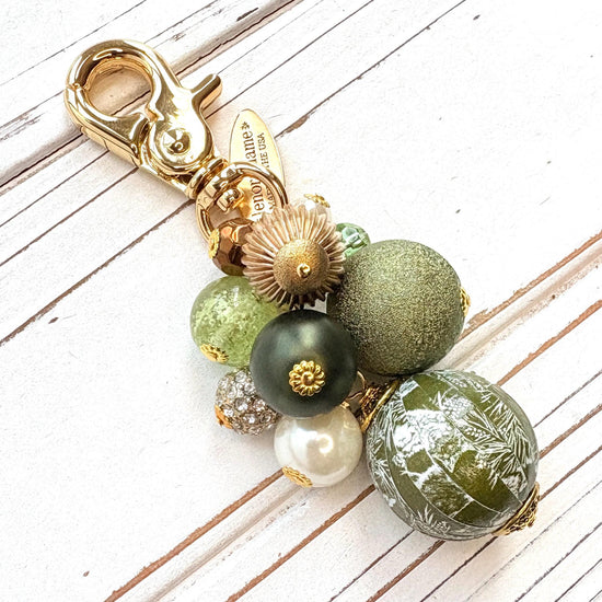 Beaded bag charm with a mix of bead styles in shades of green, gold, and pearl. One larger round bead in a army green and white toile print hangs at the bottom and a gold trigger clasp on top of all of the beads. Brass oval hang tag near the trigger clasp that says "Lenora Dame Handmade in USA". Lenora Dame bag charm.