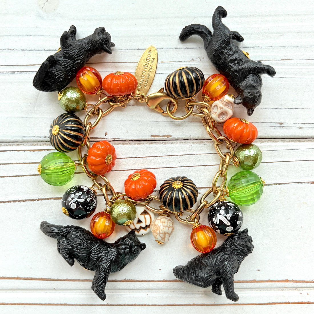 A gold chain bracelet with black cat charms, green and orange beads, and a white pumpkin charm. The bracelet has a tag that reads &
