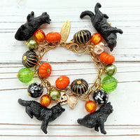 A gold chain bracelet with black cat charms, green and orange beads, and a white pumpkin charm. The bracelet has a tag that reads 'Lenora Dame' and 'Made in USA'.