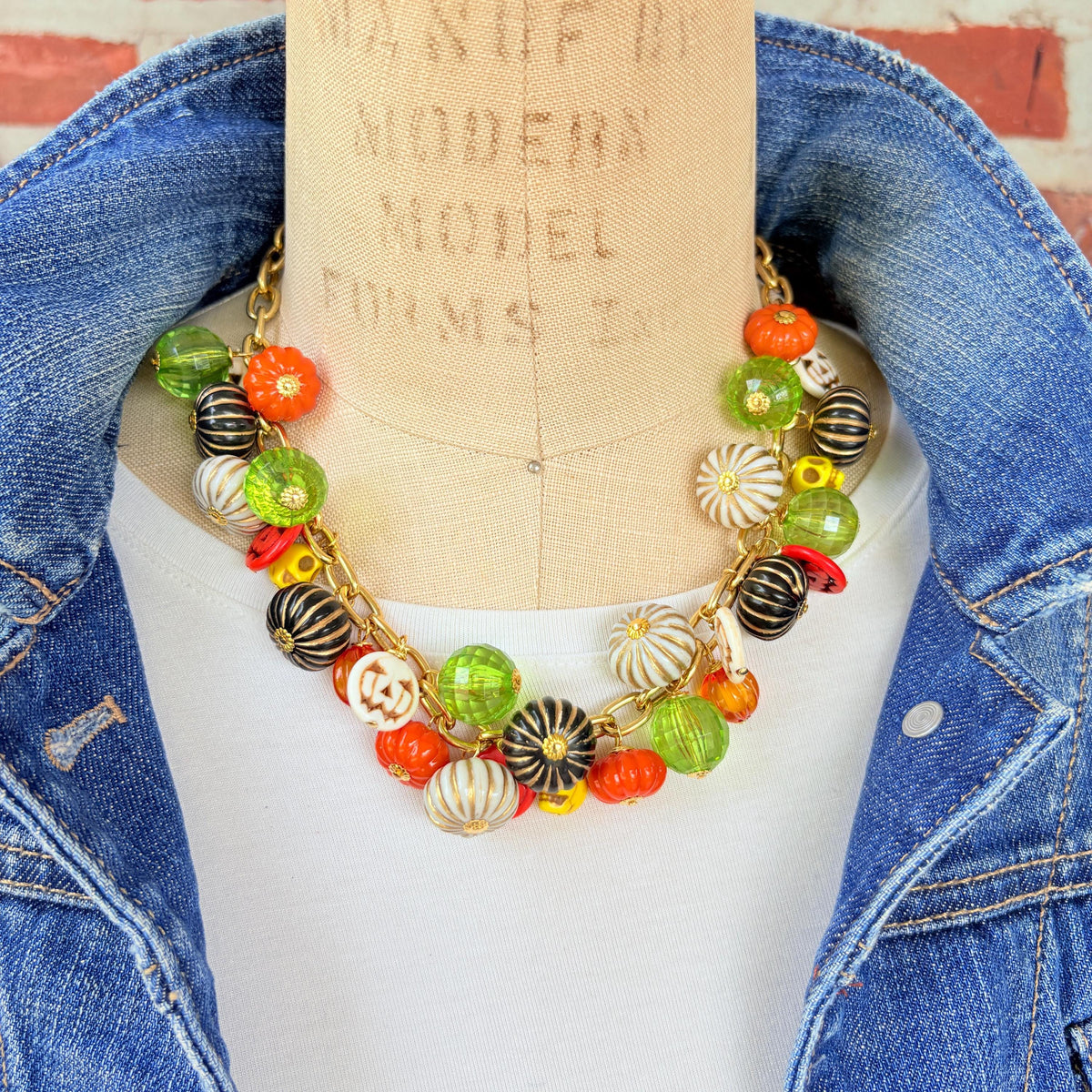 Lenora Dame necklace with a gold chain filled with orange and white jack o lantern beads, orange pumpkins, yellow sugar skull beads, black pumpkins, and green beads. Cute necklace for fall. Necklace is shown on a mannequin wearing a white t-shirt and blue denim jacket.