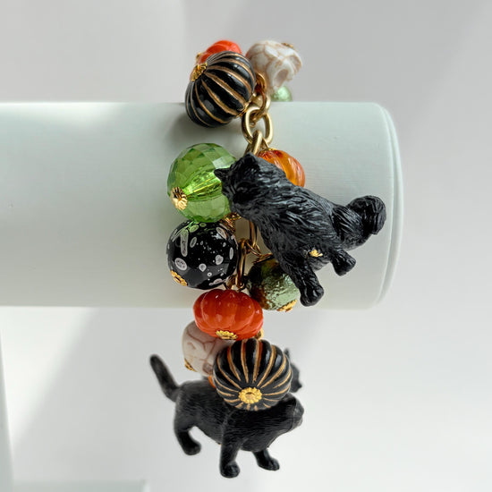 A gold chain bracelet with black cat charms, green and orange beads, and a white pumpkin charm. The bracelet has a tag that reads 'Lenora Dame' and 'Made in USA'.