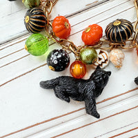 A gold chain bracelet with black cat charms, green and orange beads, and a white pumpkin charm. The bracelet has a tag that reads 'Lenora Dame' and 'Made in USA'.