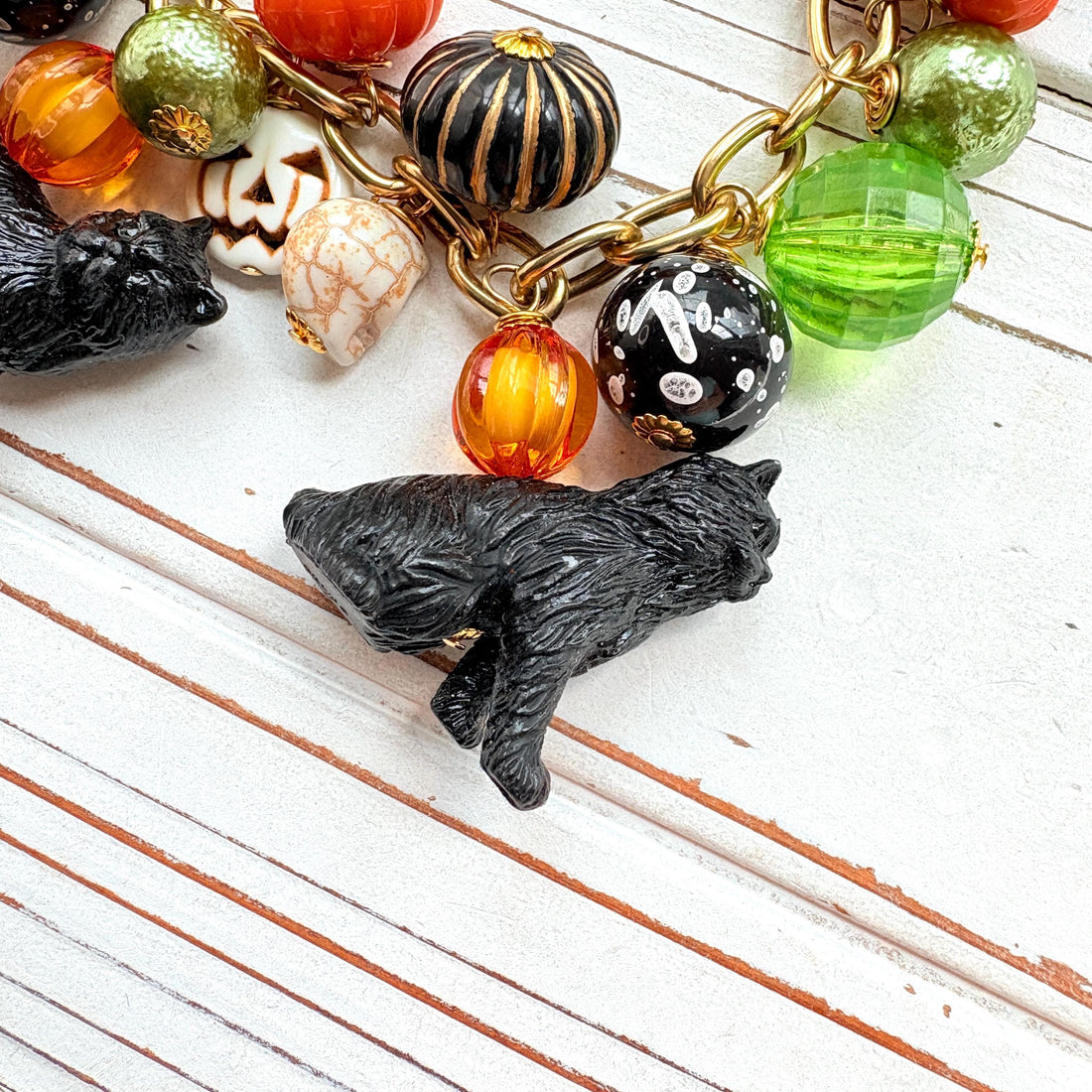 A gold chain bracelet with black cat charms, green and orange beads, and a white pumpkin charm. The bracelet has a tag that reads &