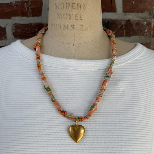 May include: A necklace with a peach-colored fabric cord, adorned with floral patterns in green, blue, and yellow. A large, gold-colored heart pendant hangs from the center, creating a vintage-inspired accessory.