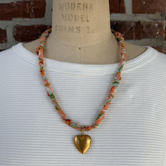 May include: A necklace with a peach-colored fabric cord, adorned with floral patterns in green, blue, and yellow. A large, gold-colored heart pendant hangs from the center, creating a vintage-inspired accessory.