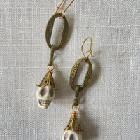 Lenora Dame Shackled Skull Earrings with white carved sugar skulls topped with brass filigree cone bead dangling from oval etched metal pieces in an antique bronze finish. Earrings hang from gold French hook ear wires. 