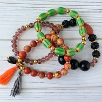 Lenora Dame 4-Piece Halloween beaded stretch bracelet set. Top set has oblong green glass beads with gold spacers. Second bracelet has orange beads with gold spacers, an orange thread tassel and a black and white thread tassel and a black tear drop bead adornment dangling from the left. The third bracelet has purple faceted beads. The last bracelet has a mix of black bumpy beads, purple faceted beads, larger round black beads and an orange faceted semiprecious stone.